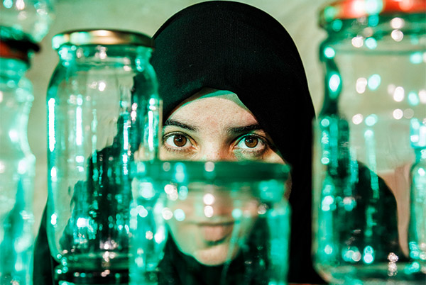 A woman from Syria with a black headscarf on looking towards the camera with a wall of jars