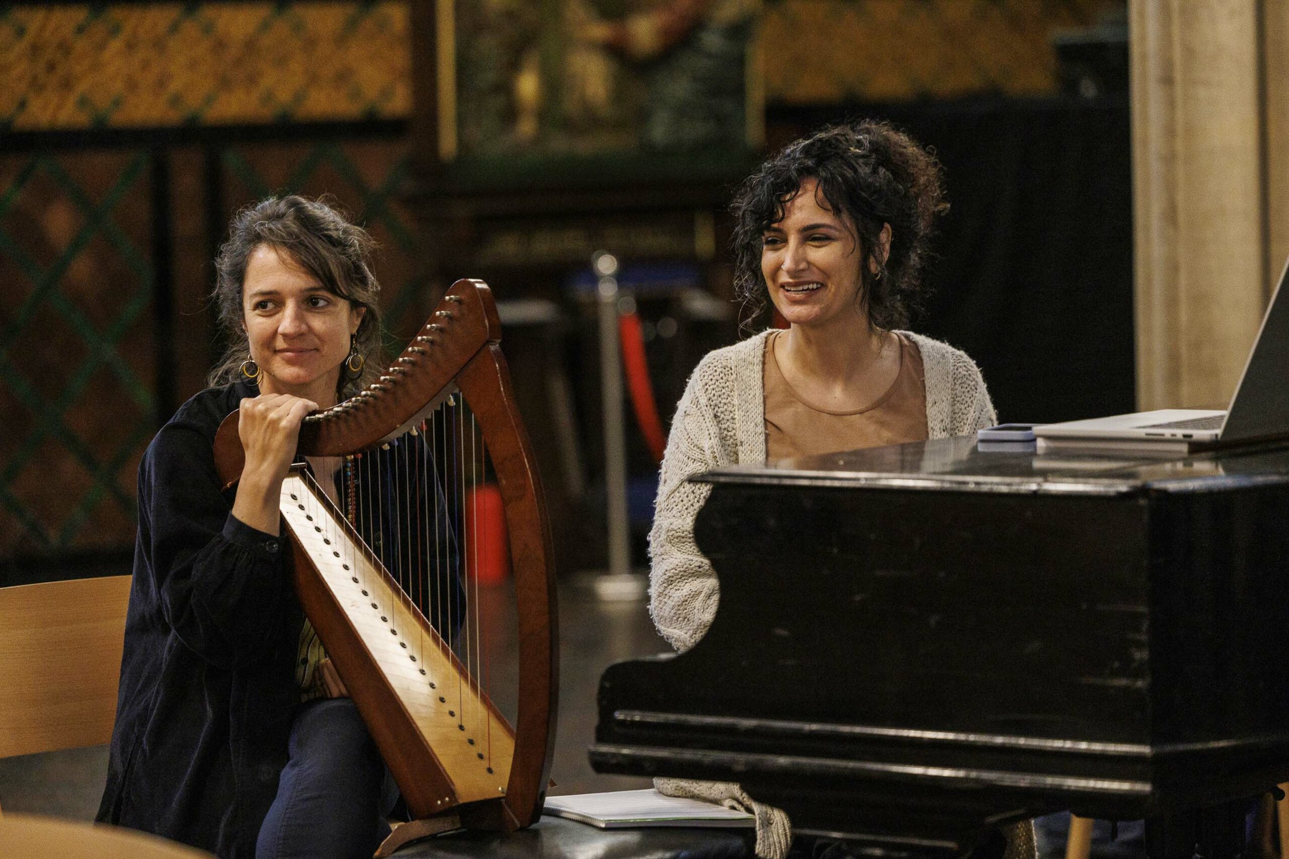 A woman sitting in front of piano and the other woman holding harp next to her